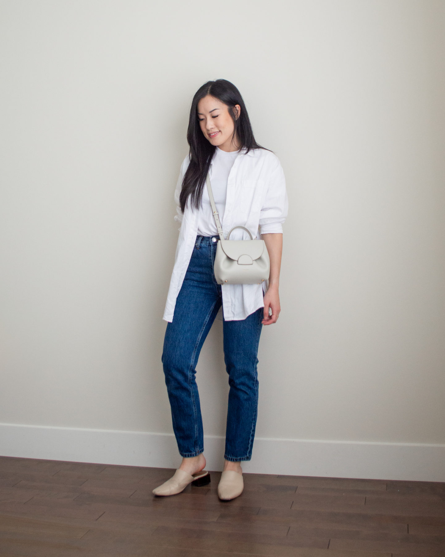 Sharon of Her Simple Sole is showcasing an outfit from her September outfit roundup, featuring a basic white T-Shirt, an oversized white button-up shirt, dark blue straight leg jeans, an off-white crossbody bag and nude mules | minimalist outfits | simple outfit ideas