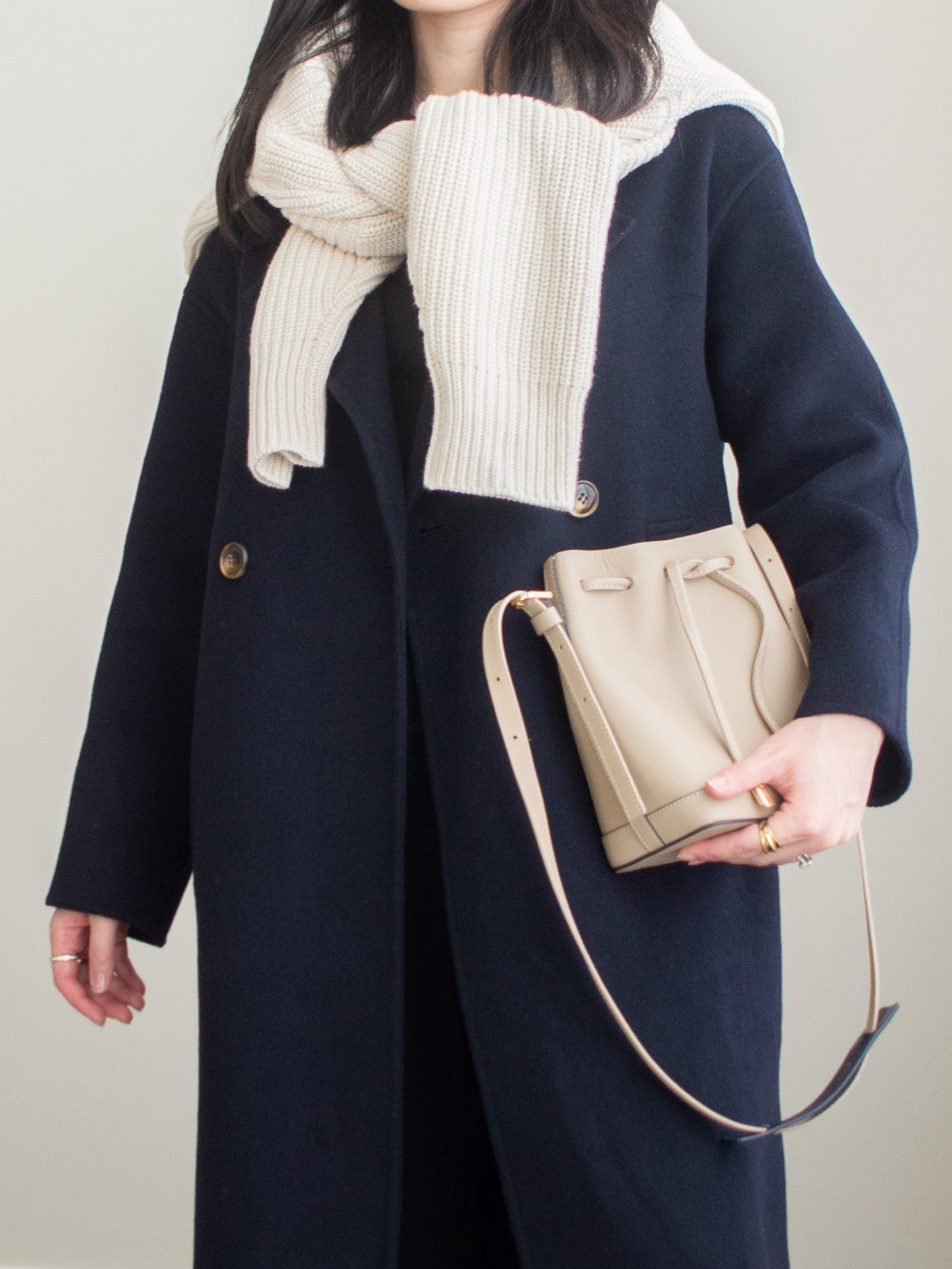 Close-up detailed outfit shot of Sharon of Her Simple Sole wearing a navy cashmere coat, cream cotton cardigan over the shoulders, black long sleeve top, black cropped trousers, taupe bucket bag and gold chunky ring | minimal style | navy cashmere coat look