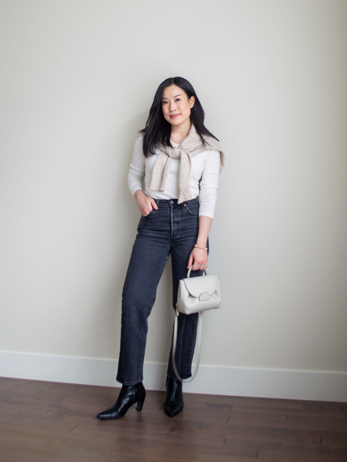 Sharon of Her Simple Sole is showcasing an outfit from a weekend trip in a backpack featuring a white long sleeve shirt, a beige cashmere sweater over the shoulders, grey straight leg jeans, black pointed toe ankle boots, and an off-white crossbody bag | minimalist outfits | fall outfit ideas | simple outfit ideas